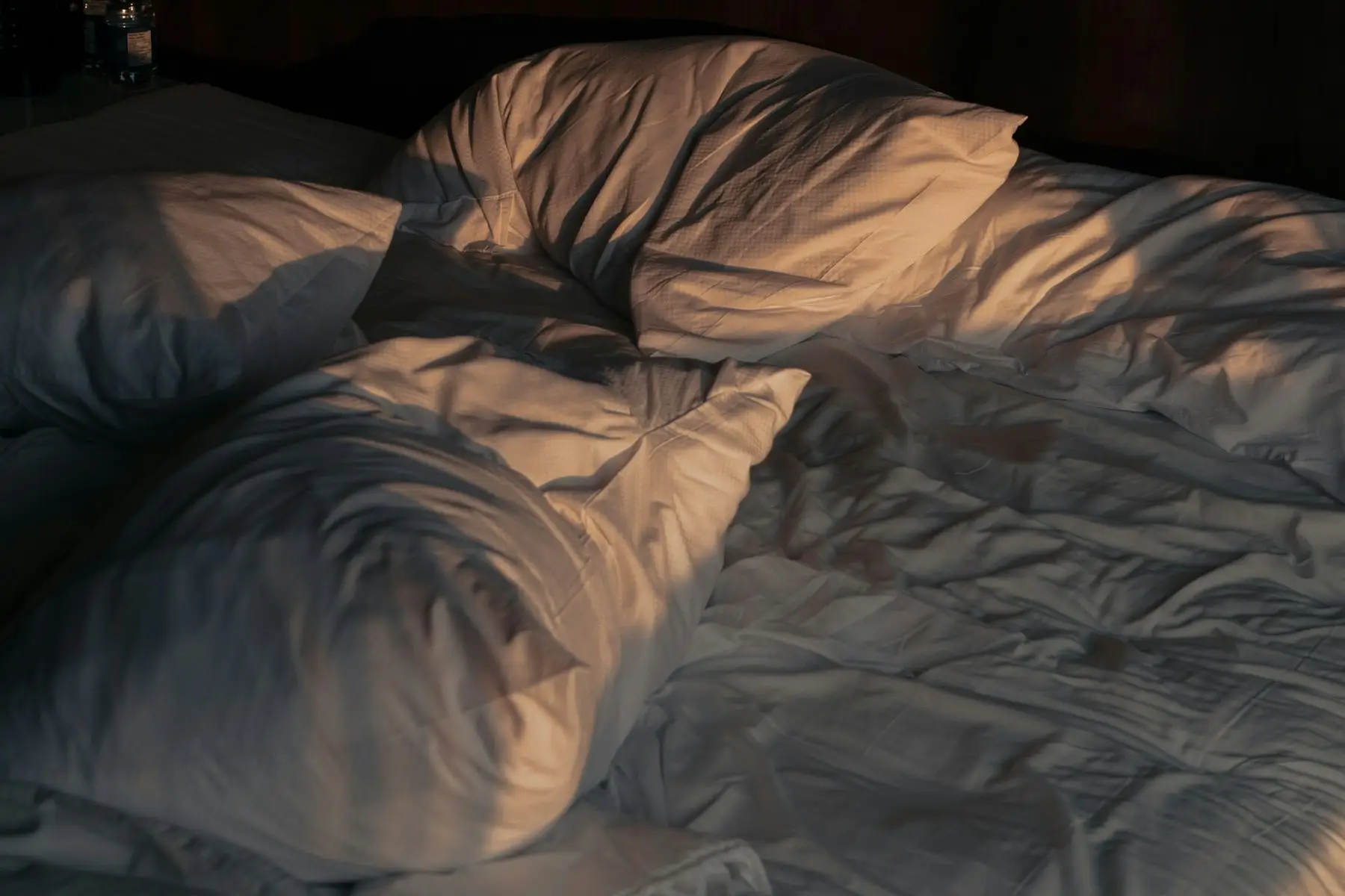 Cama desarrumada com lençóis brancos e travesseiros, numa luz suave de fim de tarde
