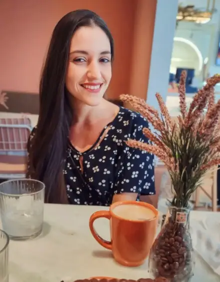 Mayara Kersbaumer sorrindo à mesa de um café, com uma caneca e um vaso de flores secas