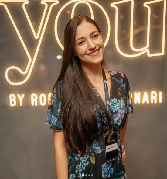 Mayara Kersbaumer sorrindo num evento, de vestido florido e crachá