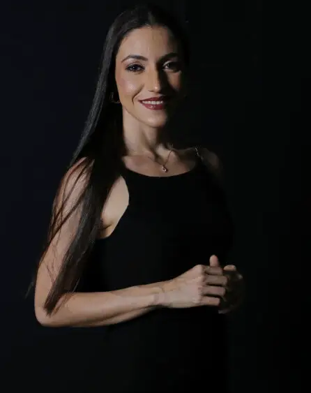 Mayara Kersbaumer sorrindo, de vestido preto, diante de um fundo escuro