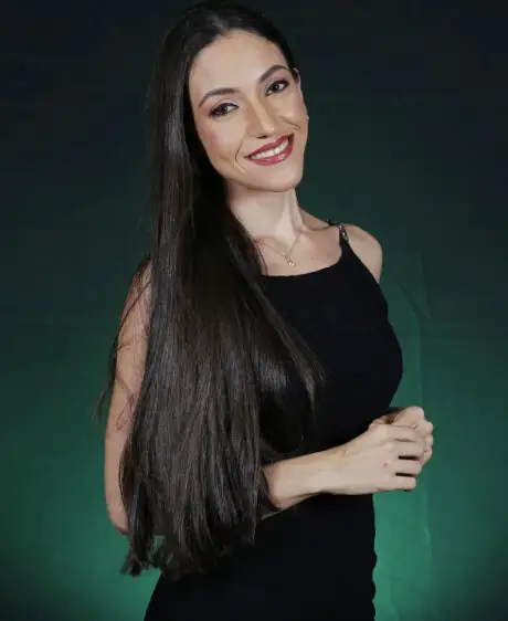 Mayara Kersbaumer sorrindo, de vestido preto e cabelo longo, diante de um fundo verde