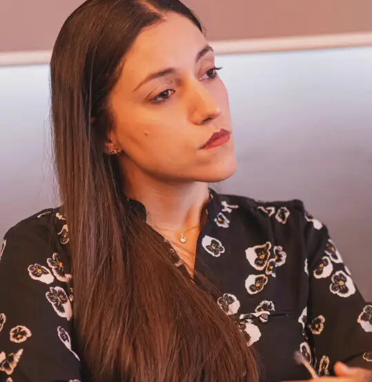 Mayara Kersbaumer, de blusa preta com flores brancas, olhando pensativa para o lado